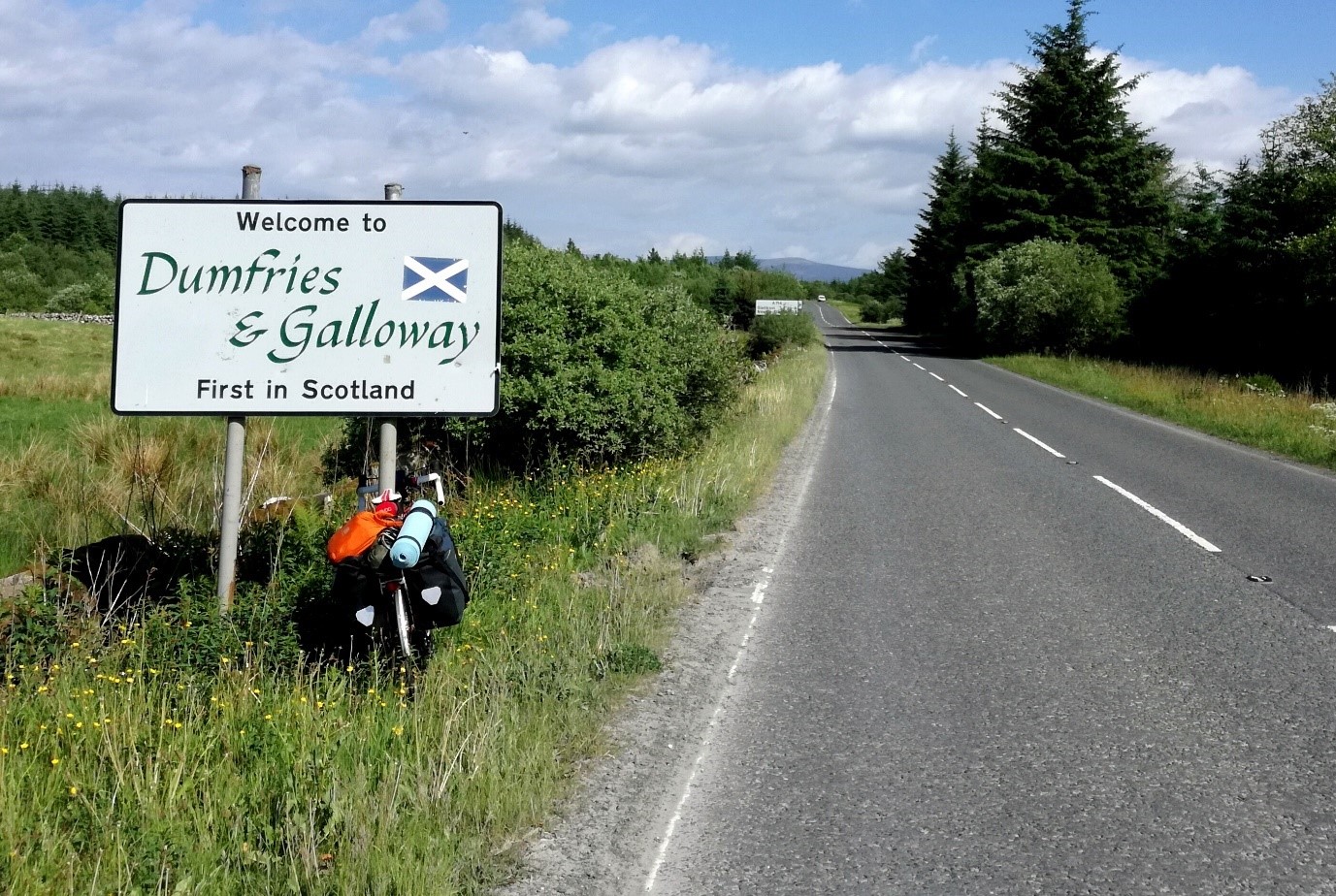 Photo of a road with sign for D&G with camping backpack resting at its foot on a sunny day